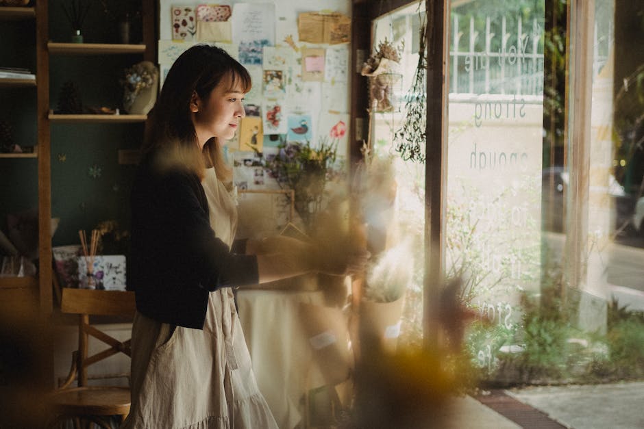 A young woman with shoulder-length brown hair, wearing a beige dress and a black cardigan, stands inside a flower shop near a large window. She is holding a bouquet of fresh flowers and appears to be 