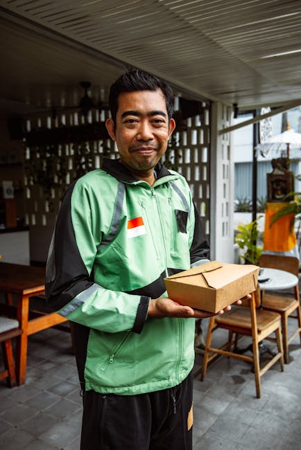 A male delivery person with short dark hair and a light brown complexion, wearing a green and black courier jacket featuring an Indonesian flag patch, standing on a covered outdoor patio. He is holdin