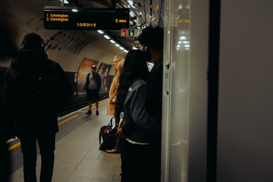 A group of commuters standing on the platform of an underground train station, with some facing the track and others waiting near the wall. The platform has a curved ceiling with illuminated lights, a