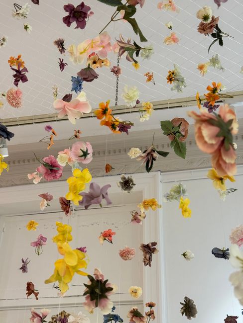 A ceiling installation of suspended artificial flowers in various colors including pink, yellow, purple, orange, white, and dark red, arranged in a scattered, floating pattern. The flowers feature det