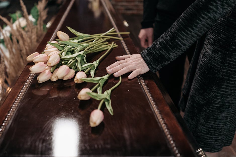 A person dressed in a black textured coat is placing a bouquet of pale pink tulips with green stems and leaves onto a polished dark wooden surface, likely a casket or memorial table. The bouquet is ti