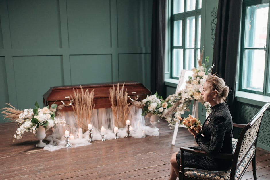 A woman with light-colored hair styled in an elegant updo, dressed in a black patterned dress, sits in a vintage armchair, holding a small bouquet of soft pink and peach flowers. She is facing a flora