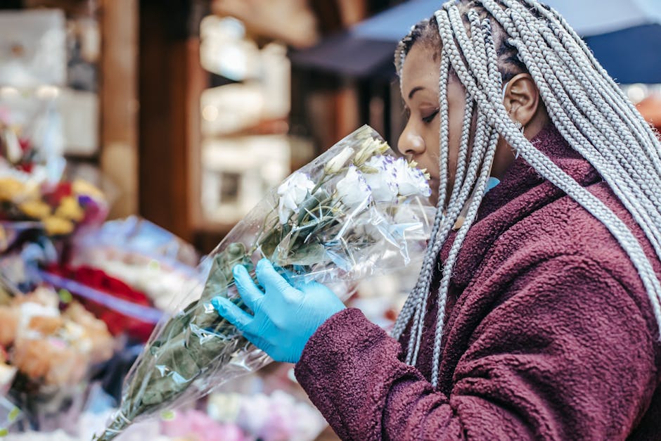 A woman with long, braided silver hair wearing a burgundy, textured fleece jacket and blue latex gloves, is closely inspecting a bouquet of white roses wrapped in clear plastic. She is holding the bou