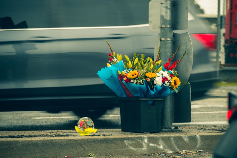 A black plastic bin filled with a vibrant floral bouquet featuring yellow sunflowers, white daisies, red roses, and green foliage, wrapped in bright blue paper with a red ribbon, is placed beside a gr
